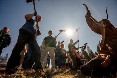 Gruppe von Menschen hält Spaten in den Händen und blickt von oben auf eine Person am Boden. Die Szene zeigt eine gemeinsame Pflanz- oder Aufforstungsaktion im Freien bei Sonnenschein und vermittelt Teamarbeit und Engagement für den Wald.