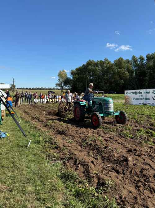 Ein Traktor pflügt ein Feld während einer landwirtschaftlichen Veranstaltung. Zuschauer, darunter Familien und Kinder, stehen am Rand des Ackers. Im Hintergrund sind Bäume, Felder und ein Banner zu sehen, darüber ein klarer blauer Himmel.