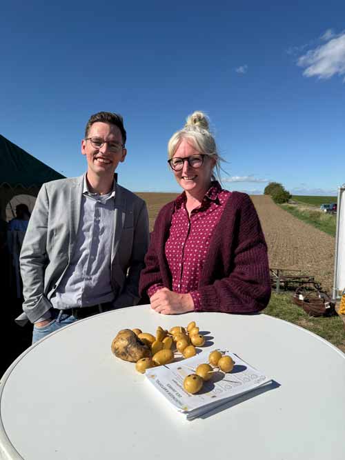 Zwei Personen stehen lächelnd an einem Stehtisch im Freien. Auf dem Tisch liegen mehrere Kartoffeln auf einem Teller. Im Hintergrund sind Felder und ein blauer Himmel zu sehen. Die Szene wirkt wie Teil einer landwirtschaftlichen oder regionalen Veranstaltung.